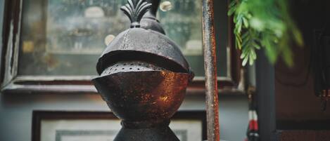 A rustic, antique metal object with a decorative top, possibly a part of a larger structure, set against a blurred background with a window and greenery.