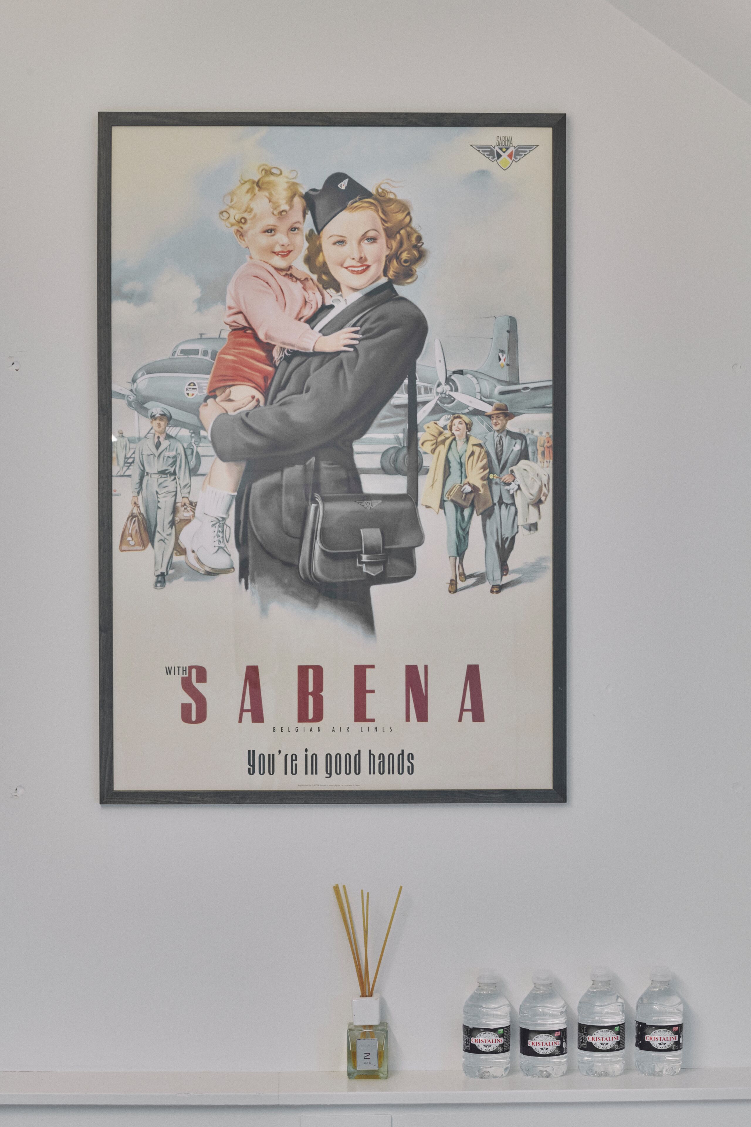 Anuncio en marco de Sabena con una mujer sosteniendo a un niño, rodeado de aviones antiguos y gente.