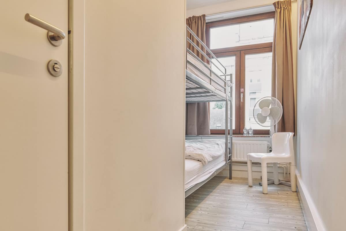 Een slaapkamer met stapelbed, raam, radiator, stoel en een deur met hendel en slot.