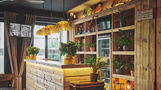 Um espaço de bar com balcão de madeira, plantas em vasos, luminárias vintage e uma placa de boas-vindas.