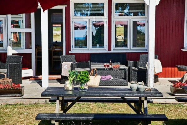Un patio al aire libre con mobiliario de mimbre, una mesa de madera con una cesta y flores, y un edificio rojo con ventanas grandes.