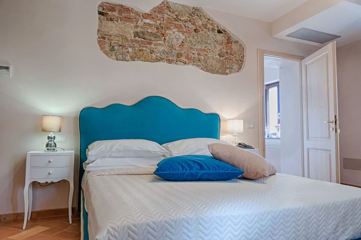 A bedroom with a blue headboard, white bedding, a small bedside table with a lamp, and a door leading to another room.