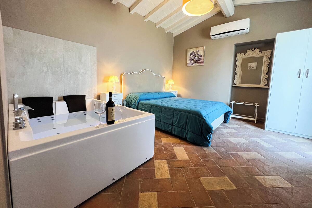 A hotel room with a large bathtub, a bed with a blue bedspread, a nightstand with a lamp, and a wardrobe.