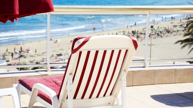 A white beach chair with red stripes and a red cushion, overlooking a beach with people and umbrellas.