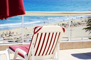 Una silla de playa blanca con rayas rojas y un cojín rojo, con vistas a una playa con gente y sombrillas.
