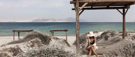 Escena de playa con una estructura de madera, un banco y una persona sentada sobre una duna de arena.