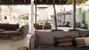 A modern lounge area with a thatched roof, wooden furniture, and a large tree providing shade.