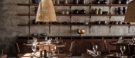 A dining area with wooden tables and chairs, shelves with pottery and kitchenware, and a hanging light fixture.