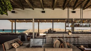 A spacious lounge area with a large window overlooking the ocean, furnished with sofas and a bar area with shelves of bottles.
