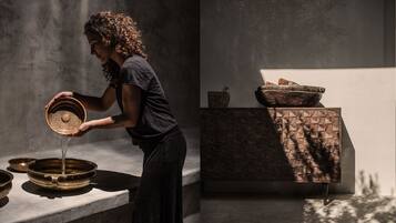 Una persona vierte agua de un cuenco a una palangana. Hay dos cuencos sobre una mesa, uno con tapa y otro sin. La habitación tiene una pared con textura y un suelo estampado.