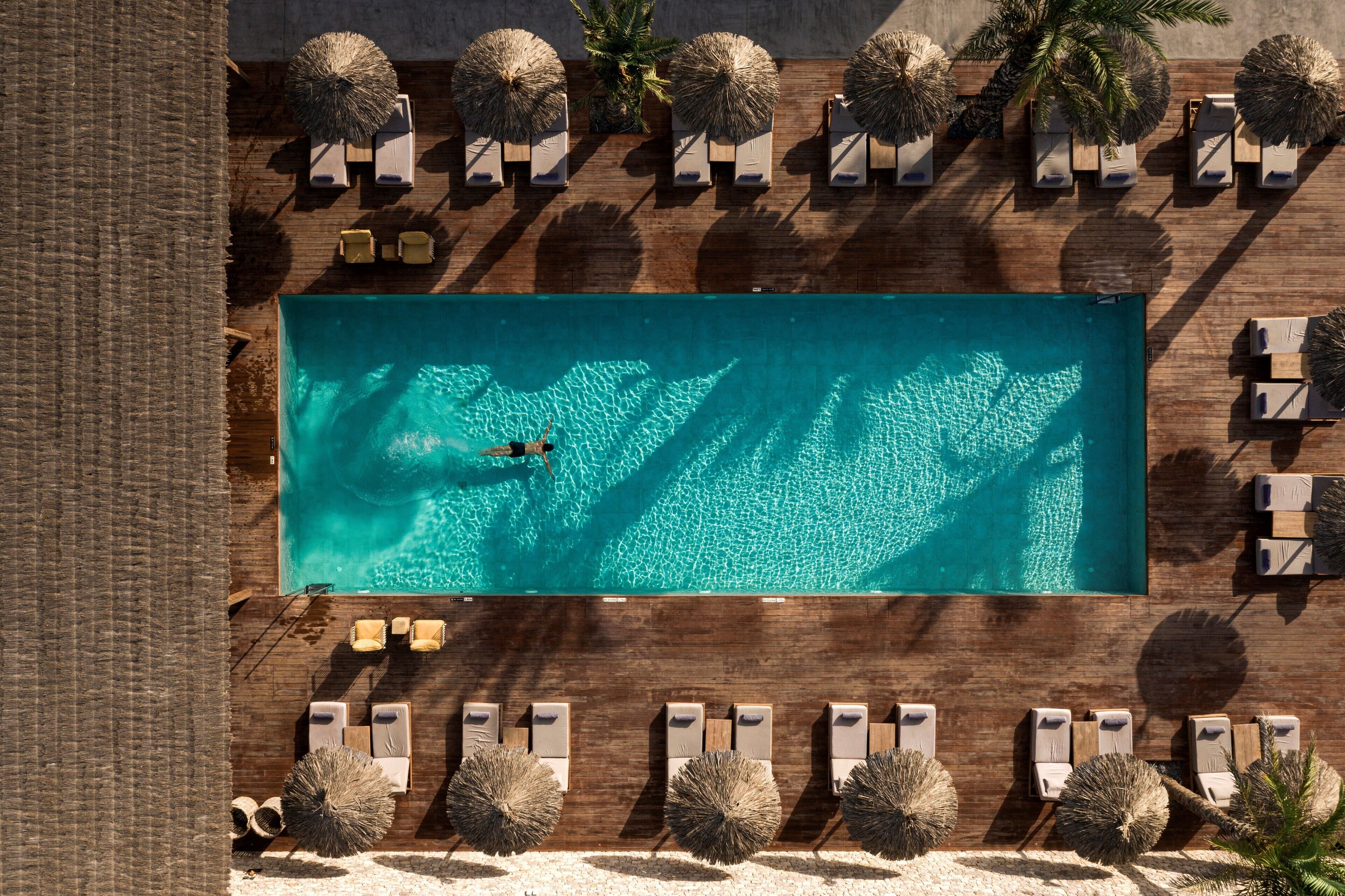 En vy från ovan av en rektangulär swimmingpool omgiven av solstolar och parasoller, med en sandstrand i förgrunden.