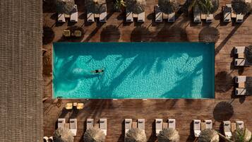 Vista aérea de una piscina rectangular rodeada de sillas de descanso y sombrillas, con una playa arenosa al frente.