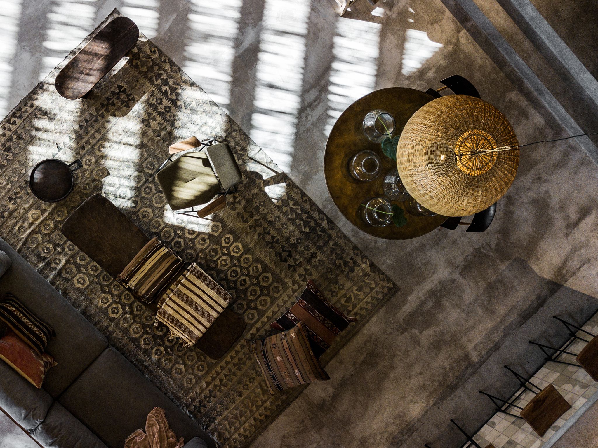 A spacious room with a large window, a round table with a woven lamp, and a patterned rug.