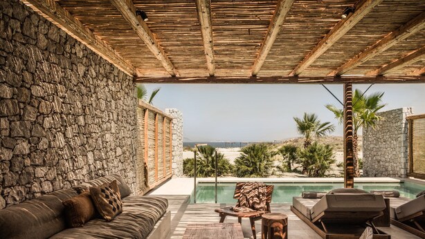 A covered outdoor seating area with a stone wall, a pool, and a view of the beach.