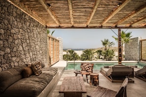 A covered outdoor seating area with a stone wall, a pool, and a view of the beach. - OKU Hotel Kos - Adults Only (Kos)