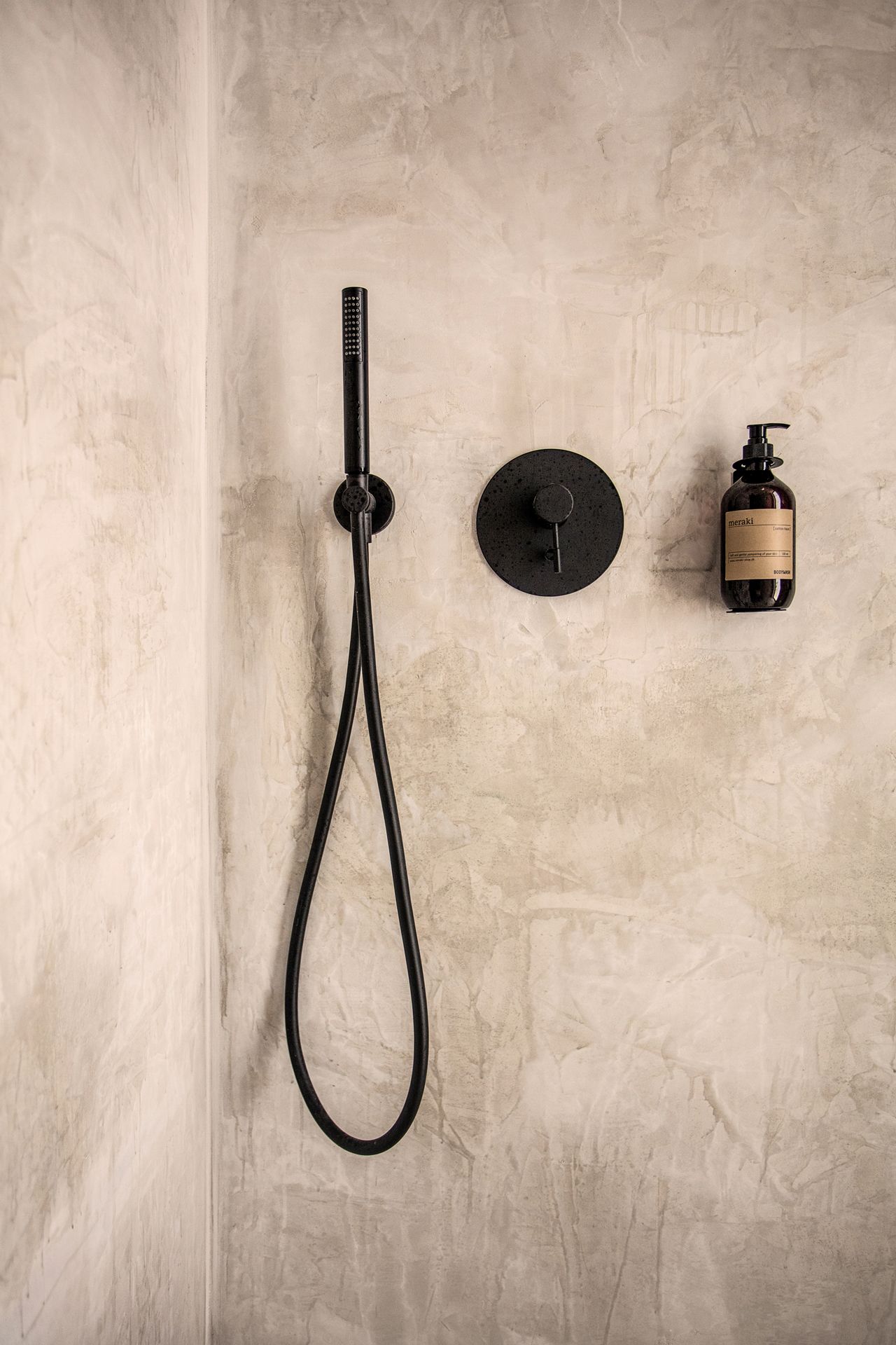 A shower with a black hose and a round black control panel, and a bottle of meraki body wash on the wall.