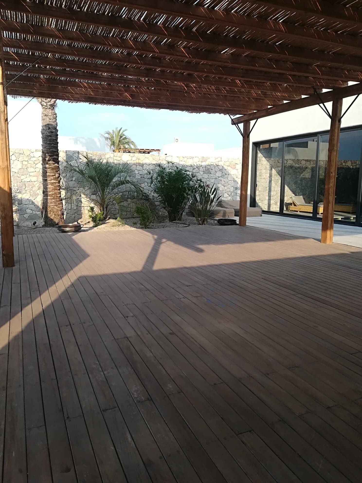 An outdoor wooden deck with a pergola, a stone wall, and a palm tree.