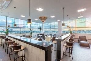 Un área de bar moderna con techo alto, ventanas amplias y vistas a la ciudad.