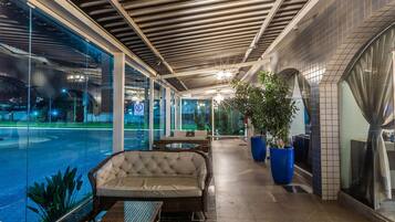 Área de estar coberta ao ar livre com vista para a piscina, com móveis de vime e plantas em vasos.