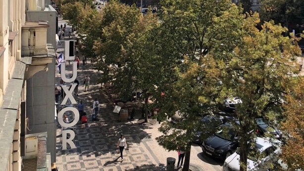 Una plaza urbana con un edificio grande al fondo, una calle bordeada de árboles y un letrero que dice 'LUXOR'.