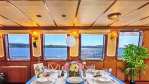 Un coin repas avec une table dressée pour quatre personnes, de grandes fenêtres donnant sur l’eau et une plante en pot placée à droite.
