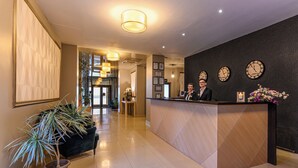 Un espace d’accueil d’hôtel comprenant un comptoir de réception, deux réceptionnistes, une plante en pot et un mur orné d’horloges.