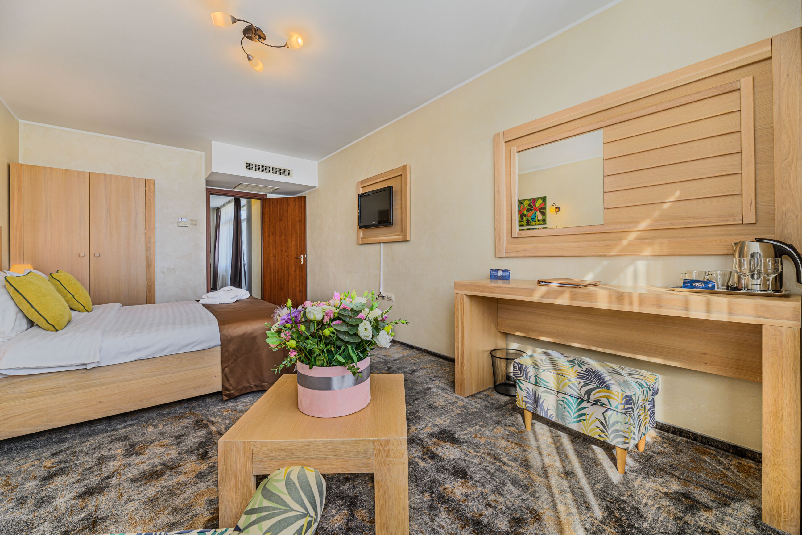 A hotel room with a bed, a small table with a flower vase, a desk with a mirror, and a sofa.