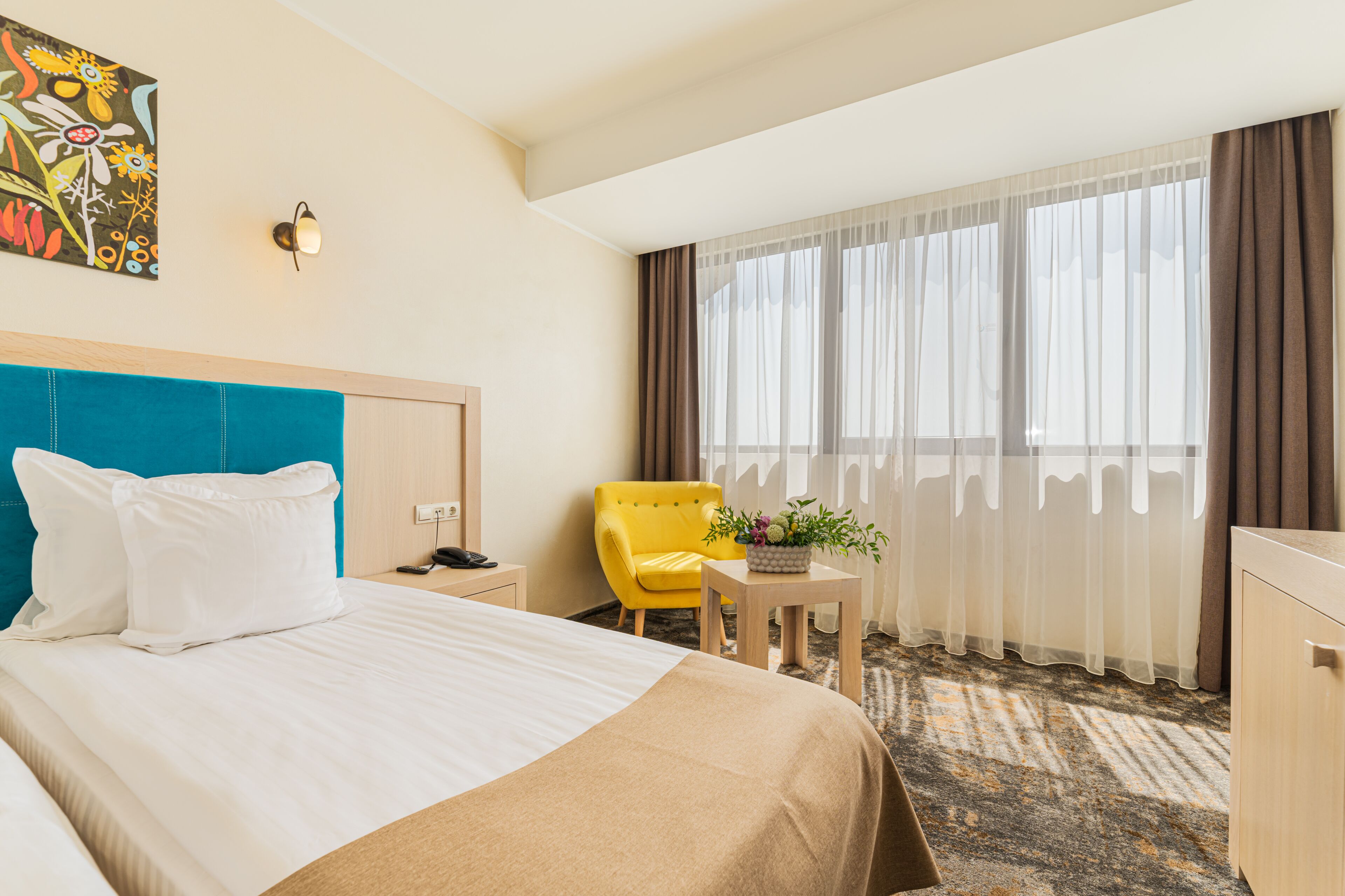 A hotel room with a bed, a yellow armchair, a small table with a plant, a desk with a lamp, and a painting on the wall.