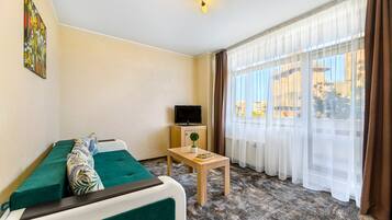 Habitación de hotel con un sofá verde, una mesita con una planta, un televisor y un ventanal con cortinas.