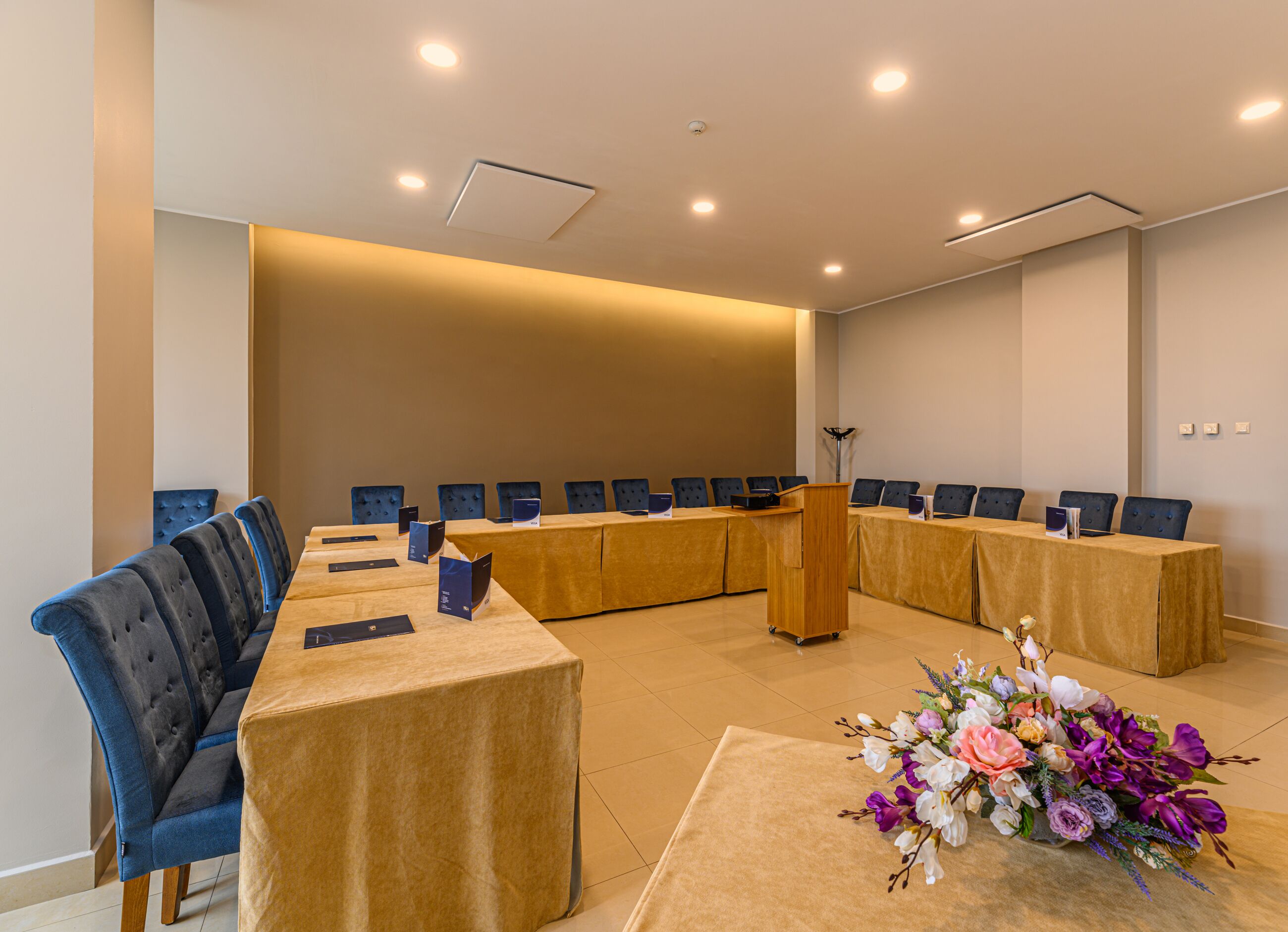 Una sala de conferències amb una taula llarga, cadires blaves, un escenari i un arranjament floral.