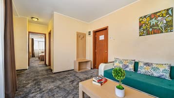 Habitación de hotel con un sofá verde, una pequeña mesa con una planta y libros, una puerta de madera y un pasillo que conduce a otra habitación.