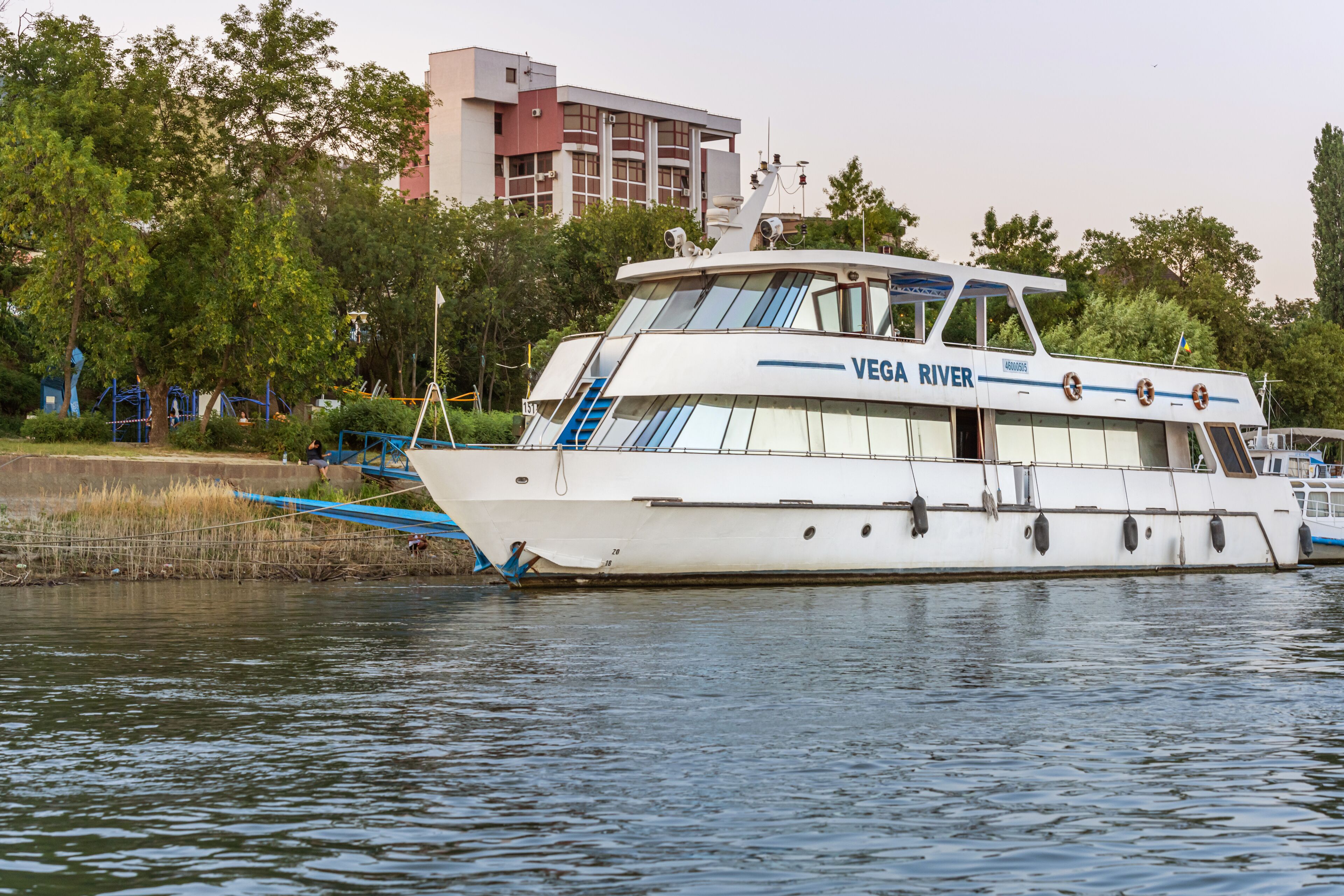 A white boat named "VEGA RIVER" docked near a park with playgrounds and trees.