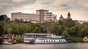 Une vue sur le fleuve, avec un grand bâtiment portant l’inscription « Paleza Hotel » en arrière-plan, un bateau amarré sur la rive et une petite structure en bois sur la droite.