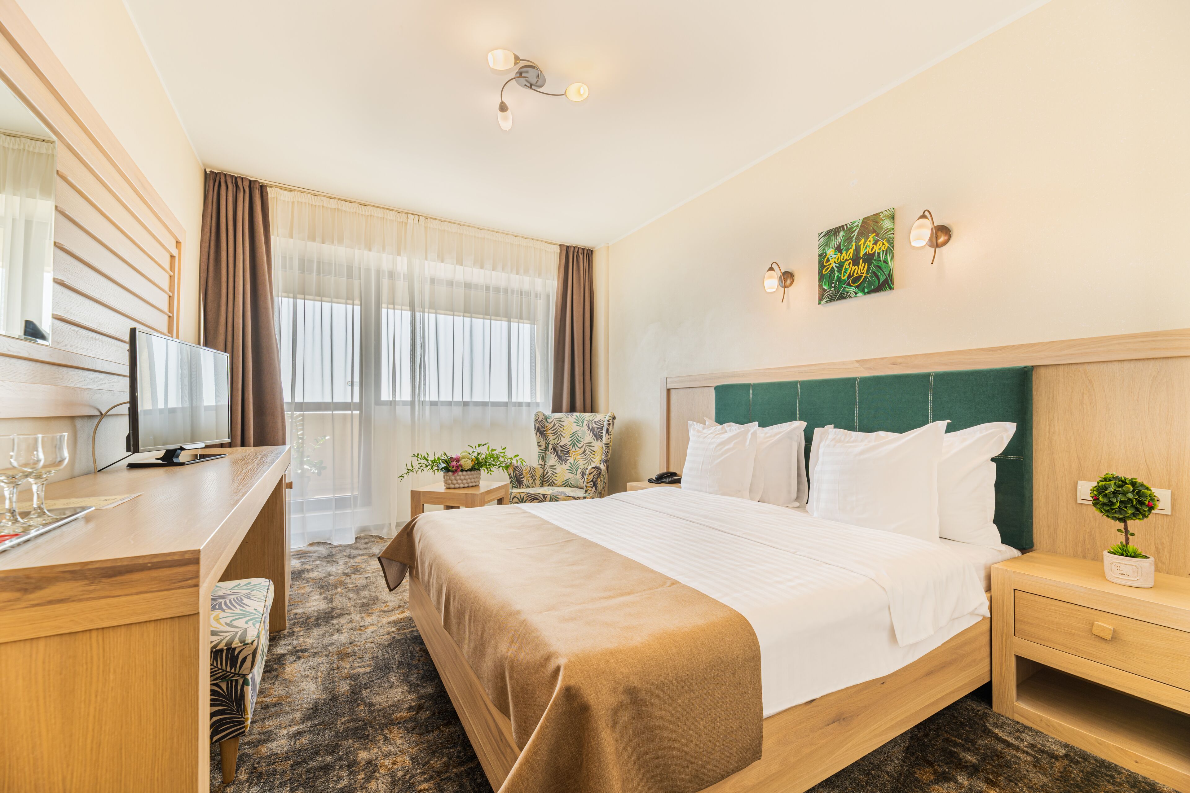 A hotel room with a large bed, a desk with a computer and glasses, a chair, and a small table with a plant.