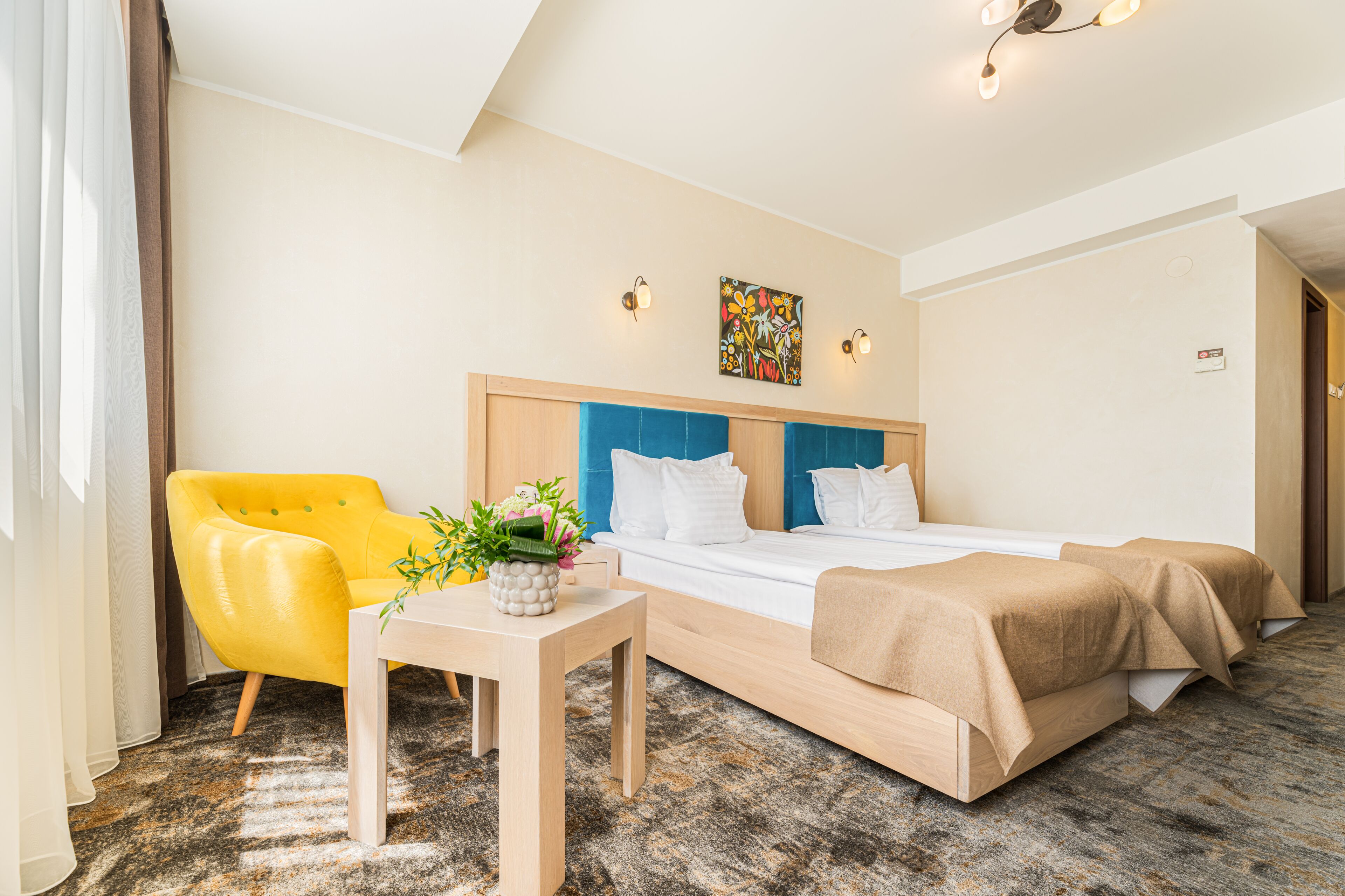 Habitación de hotel con cama, un sillón amarillo, una mesita con una planta y un cuadro en la pared.