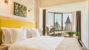Une chambre d’hôtel avec un grand lit, une tête de lit, une fenêtre donnant sur des immeubles, une chaise et une plante.
