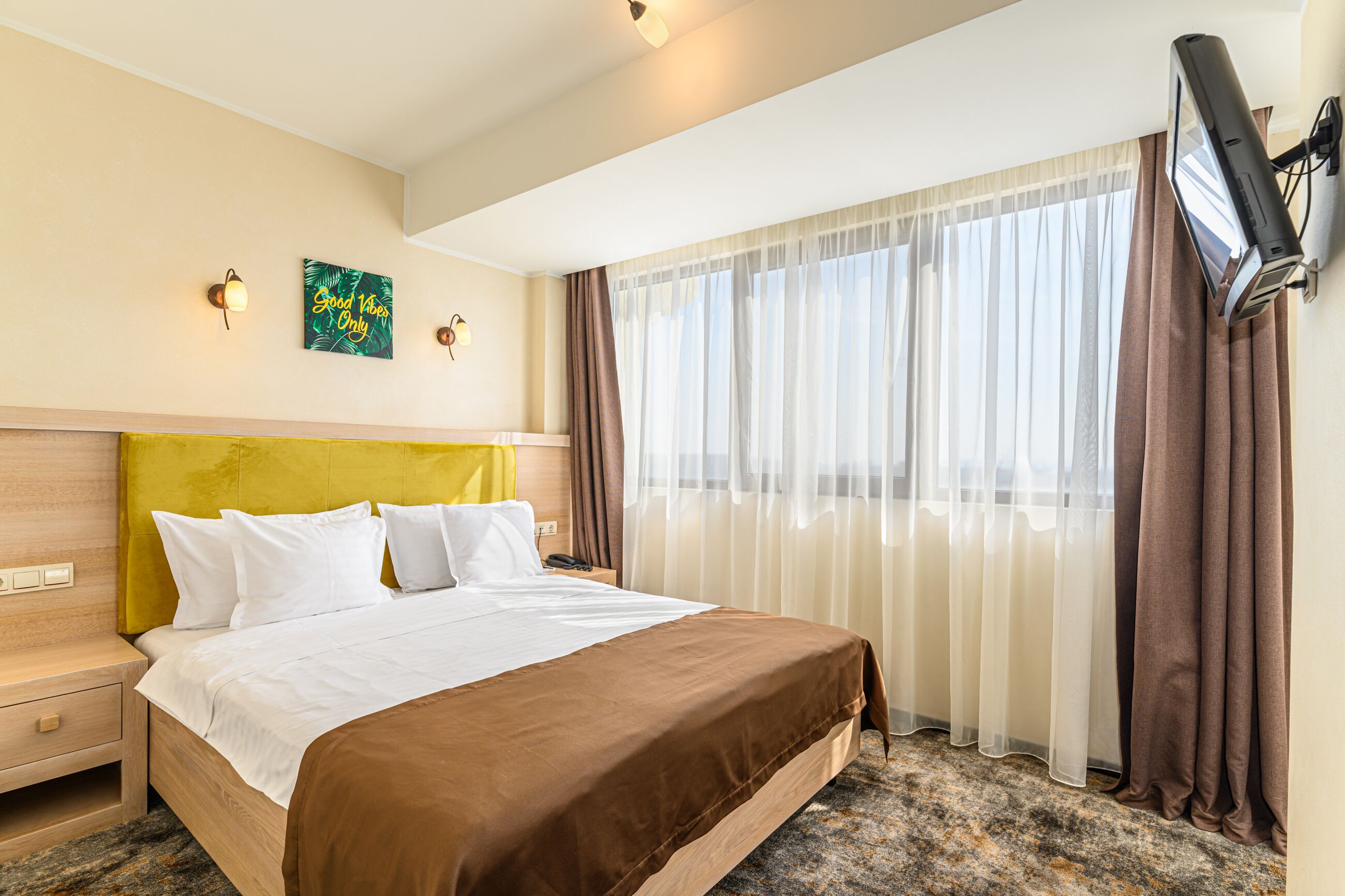 A hotel room with a bed, a nightstand, a wall-mounted TV, and a window with sheer curtains.
