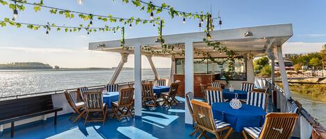 A covered outdoor seating area with tables and chairs, overlooking a body of water.
