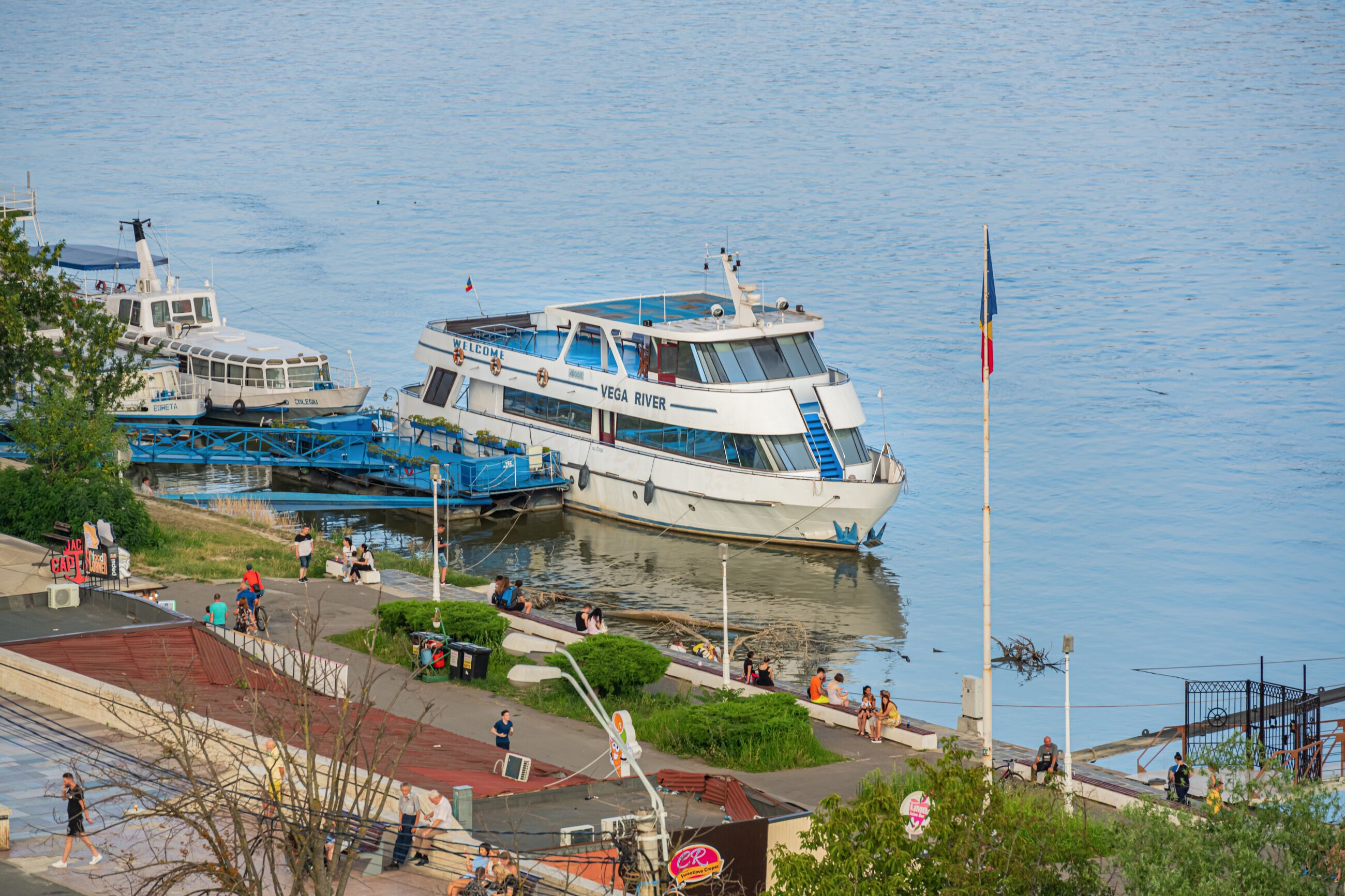 A riverfront area with a docked boat named "VEGA RIVER" and a pedestrian walkway alongside.