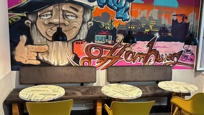 Un área de estar con un mural, mesas con manteles individuales estampados con diseño de periódico y sillas amarillas.