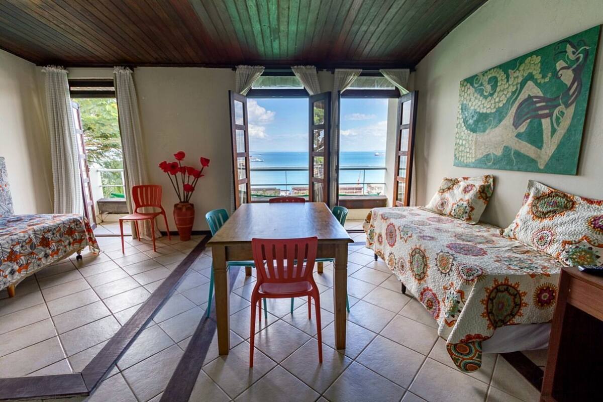 A room with a wooden table, two chairs, a bed with patterned bedding, and a view of the sea through open windows.