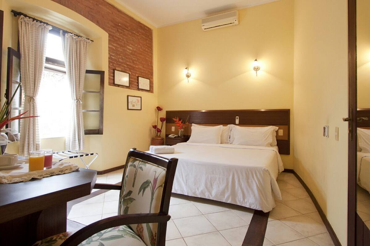 A hotel room with a bed, bedside table, chair, and a window with curtains.