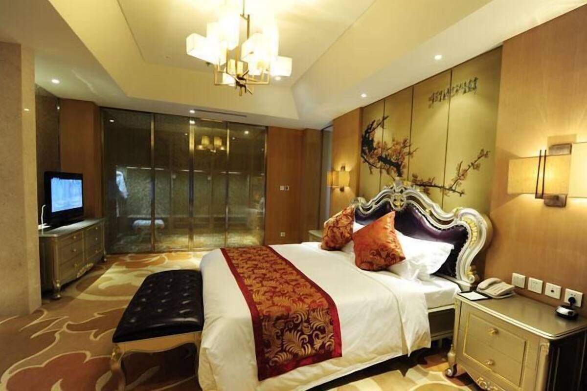 A hotel room with a large bed, a television, a desk with a telephone, and a wall-mounted light.