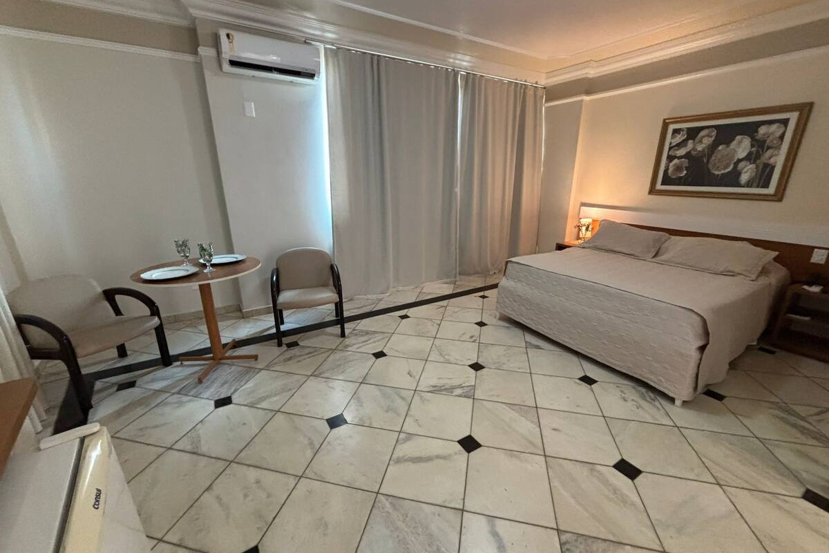 A hotel room with a bed, a small table with chairs, an air conditioning unit, and a painting on the wall.