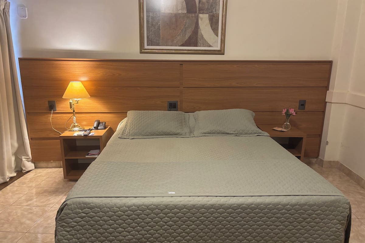 A neatly made bed with a quilted cover, a bedside lamp, a phone, and a small table with a vase of flowers.
