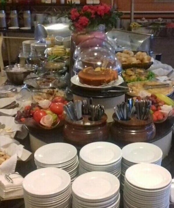 A buffet setup with various food items, including fruits, vegetables, and a cake, arranged on a table with stacked plates and serving utensils. - Tamareiras Park Hotel (Uberaba)