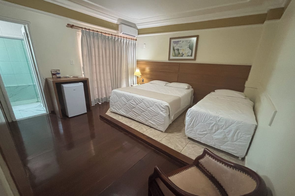 A hotel room with two beds, a desk, a chair, a small fridge, and a window with curtains.