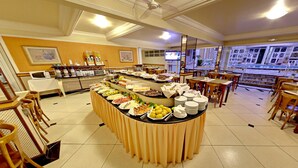 A buffet setup with various food items and a television screen in a well-lit room with tiled flooring and wooden chairs. - Tamareiras Park Hotel (Uberaba)