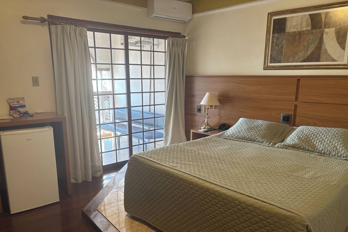 A hotel room with a bed, a nightstand, a lamp, a desk with a refrigerator, a window with curtains, and a painting on the wall.