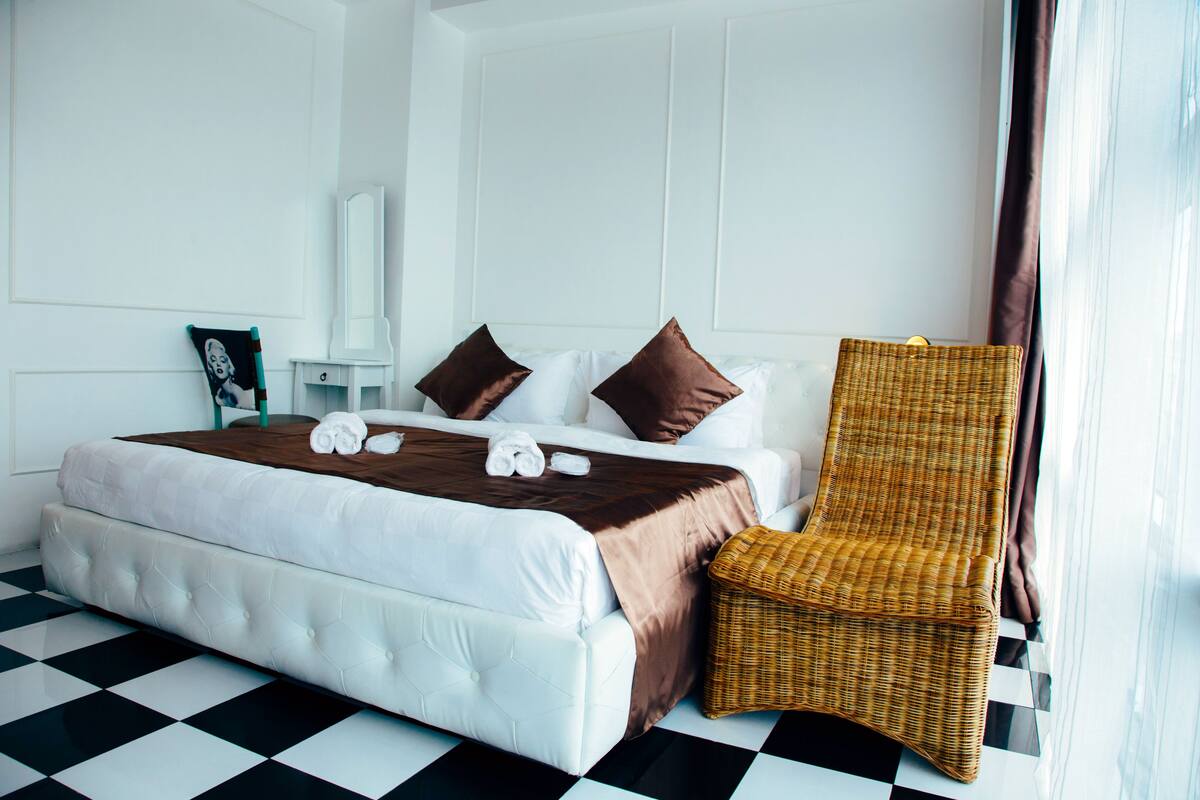 Ein Hotelzimmer mit Schachbrettmuster-Boden, einem großen Bett mit weißer und brauner Bettwäsche, einem Korbstuhl und einem weißen Nachttisch.